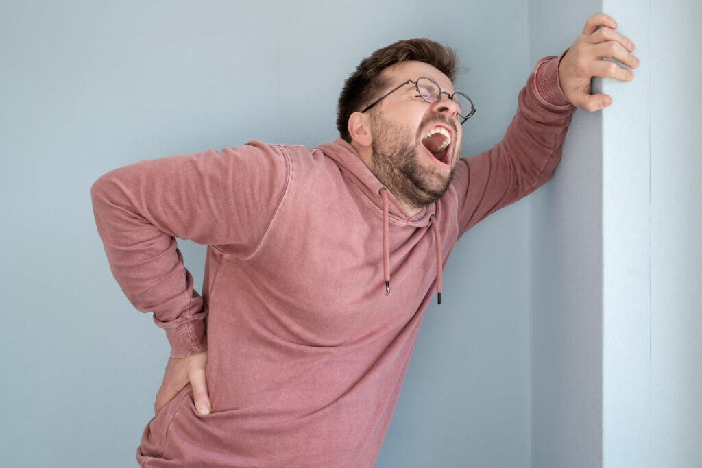 European man is suffering from severe back pain, he is screaming while holding hand against the wall